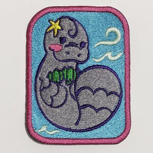 May include: Embroidered iron-on patch featuring a cartoon gray sea creature with a yellow star on its head, holding a green seaweed. The creature is surrounded by blue waves on a light blue background with a pink border.