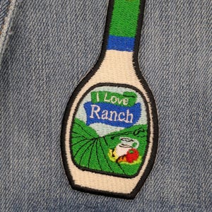 May include: Embroidered patch in the shape of a ranch dressing bottle. The patch features the text "I Love Ranch" and a graphic of a field with a tomato and a cup. The patch is outlined in black and has a white, green, and blue colour scheme.