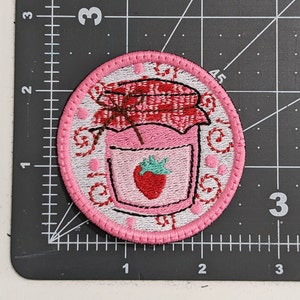 Könnte beinhalten: Ein rosa-weiß gestickter Aufnäher mit einem Glas Erdbeermarmelade. Das Glas hat einen roten Deckel und eine Erdbeere darin. Die Worte "Erdbeermarmelade" sind um das Glas herum gestickt.