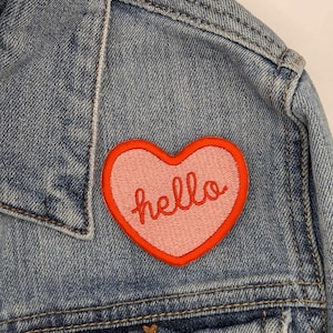 May include: A blue denim jacket with a pink heart-shaped patch that says "hello" in red embroidery.