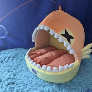 May include: A novelty pet bed shaped like an anglerfish, featuring an orange head with a black star, white teeth, and a yellow body. The bed has three orange cushions inside and a dangling blue pom-pom toy.