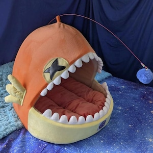 May include: Orange and yellow plush pet bed shaped like a cartoon fish with a wide open mouth and white teeth. The bed has a red interior and a dangling blue pom-pom toy. A black star is on the side.