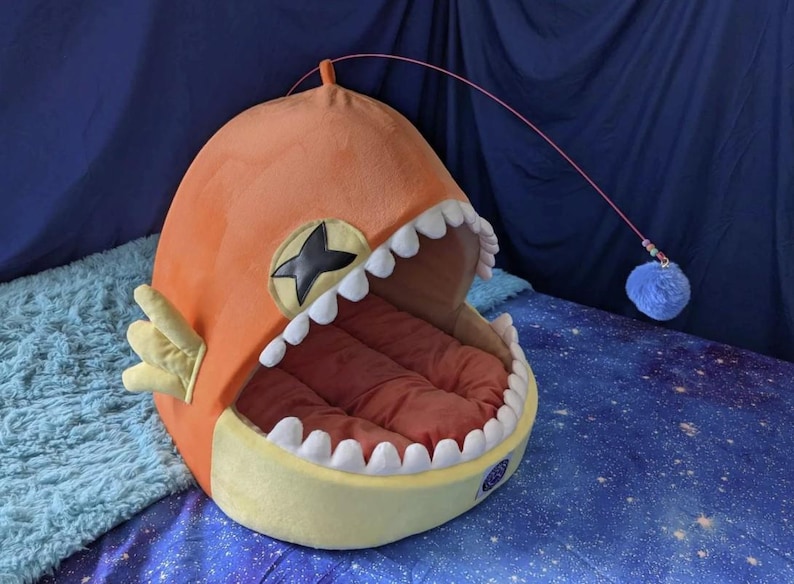 May include: Orange and yellow plush pet bed shaped like a cartoon fish with a wide open mouth and white teeth. The bed has a red interior and a dangling blue pom-pom toy. A black star is on the side.