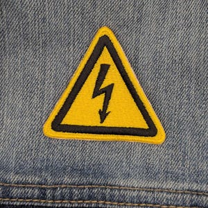 May include: Yellow and black embroidered patch in the shape of a warning triangle with a lightning bolt symbol. The patch is sewn onto a blue denim fabric.