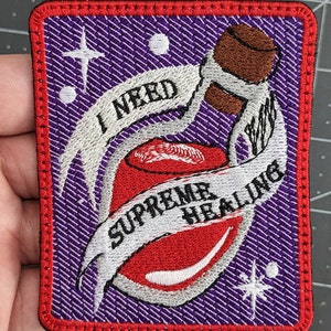 Puede incluir: Un parche bordado morado con una botella de poción roja y texto blanco que dice "Necesito curación suprema".