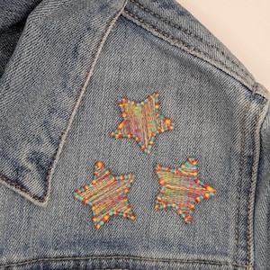 May include: A denim jacket with three embroidered stars in rainbow colors. The stars are stitched with colorful thread and are arranged in a triangular pattern.