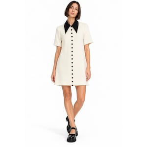 May include: A short-sleeved, off-white mini dress featuring a contrasting black pointed collar and a vertical row of small black buttons running down the center front. The dress has a structured, straight-cut silhouette that ends above the knees.