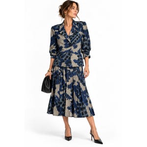 May include: A two-piece set consisting of a long-sleeved blazer with notched lapels and a matching pleated midi skirt. Both pieces feature a beige base with a dark blue and black abstract brushstroke pattern. A black clutch is held in one hand.