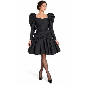 May include: A black knee-length dress featuring long sleeves with voluminous puffed shoulders, a sweetheart neckline, and a fitted bodice with diagonal pleating. The waist is accented with two small bows, leading into a full, gathered skirt.
