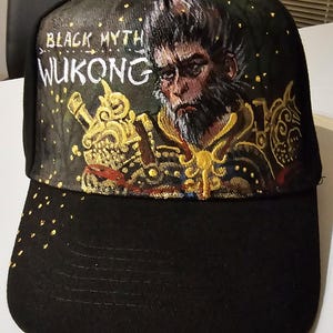 Puede incluir: Gorra de béisbol negra con un diseño pintado a mano que presenta un personaje de mono con barba y las palabras "BLACK MYTH WUKONG" en letras doradas.
