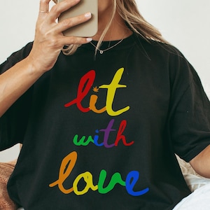 May include: Black t-shirt with the text "lit with love" in rainbow colors. The shirt is worn with light gray sweatpants and a tan beanie. The person is sitting in a wooden chair.