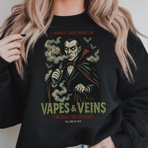 Könnte beinhalten: Schwarzes Sweatshirt mit einem Grafikdesign eines vampirischen Wesens, das dampft. Der Aufdruck enthält den Text "Vapes & Veins" und "Too High for Daylight, Halloween 1979". Der Vampir trägt einen schwarzen Anzug und einen roten Umhang.