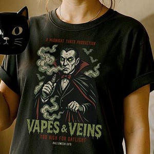 May include: Black t-shirt with a graphic of a vampire holding a vape pen, with the text "Vapes & Veins". The shirt also features the text "Too High for Daylight" and "Halloween 1979". A black cat-shaped mug is held in the hand.