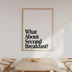 Op de afbeelding: Een eetkamer met een lichte houten tafel en stoelen. Een ingelijste print aan de muur toont de tekst "What About Second Breakfast?" in zwart. De kamer heeft een minimalistisch, modern design met natuurlijk licht.