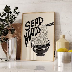 Op de afbeelding: Een ingelijst print met de tekst "Send Noods" in zwarte letters. De print toont een zwart-witte illustratie van een kom noedels.