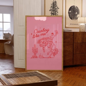 May include: Pink poster with a cowboy riding a bucking horse. The text "Cowboy like me" is written in red above the cowboy. A cactus is in the background.