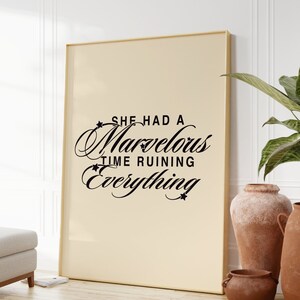 Op de afbeelding: Een beige ingelijste poster met zwarte tekst die "She had a marvelous time ruining everything" luidt. De poster hangt aan een witte muur met een plant in een pot en een houten vloer.