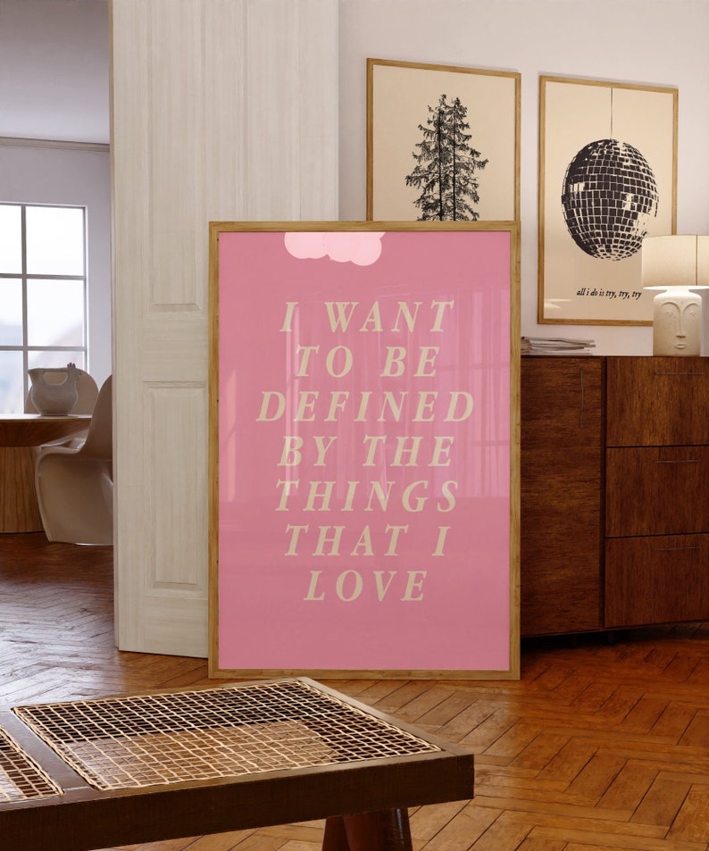 May include: A framed pink poster with the text "I WANT TO BE DEFINED BY THE THINGS THAT I LOVE" in a light beige font. The poster is in a room with other framed art and wooden furniture.