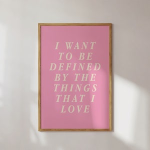 May include: A framed pink art print with the text "I WANT TO BE DEFINED BY THE THINGS THAT I LOVE" in a serif font. The frame is a light brown color, and the background is a white wall.