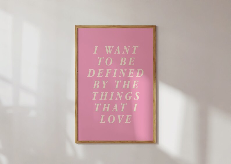 May include: A framed pink art print with the text "I WANT TO BE DEFINED BY THE THINGS THAT I LOVE" in a serif font. The frame is a light brown color, and the background is a white wall.