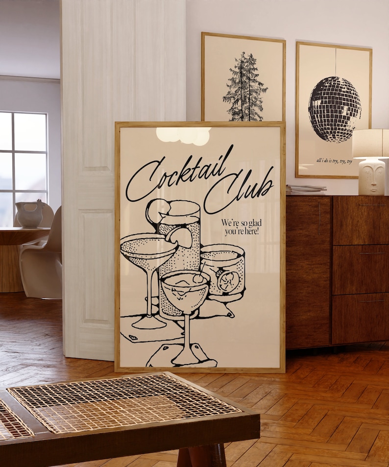 May include: A framed print with the words "Cocktail Club" and illustrations of cocktail glasses and a shaker. The print also includes the phrase "We're so glad you're here!" and is displayed in a room with other framed art.