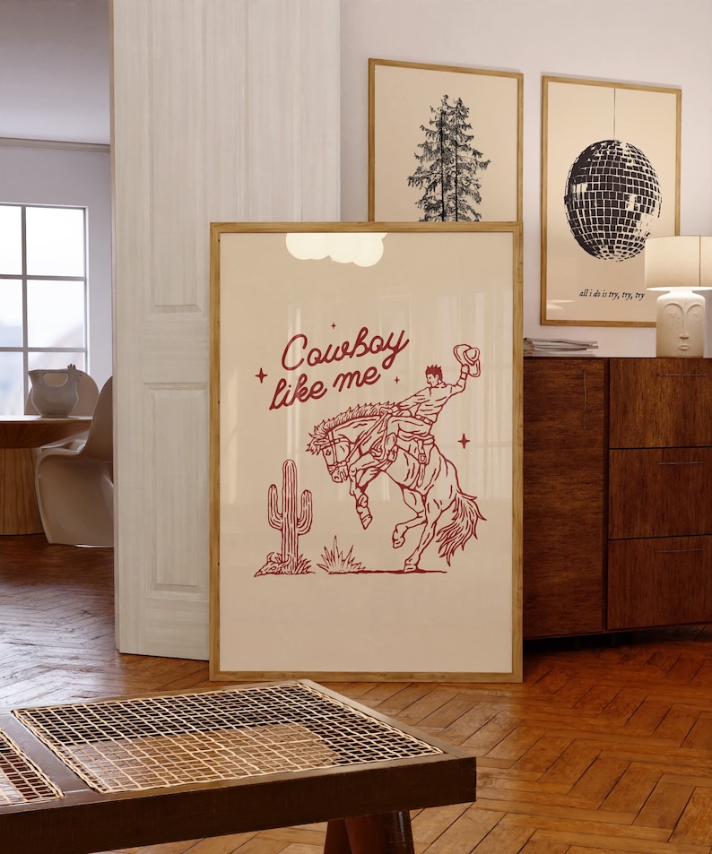 Może przedstawiać: Oprawiony wydruk z beżowym tłem przedstawia czerwoną ilustrację kowboja na koniu rodeo z napisem "Cowboy like me". Kaktus i gwiazdy są r&oacute;wnież zawarte w projekcie. Inne oprawione dzieła sztuki są widoczne w tle.