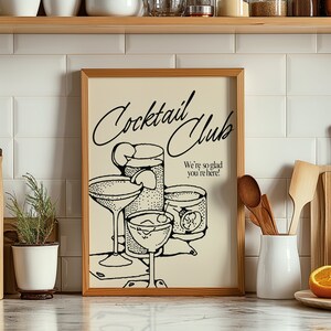 May include: A framed print with a vintage-style illustration of cocktail glasses. The print, in a light beige hue, features the words "Cocktail Club" in elegant script, along with the phrase "We're so glad you're here!" in a classic font. The artwork is set against a white tile background.