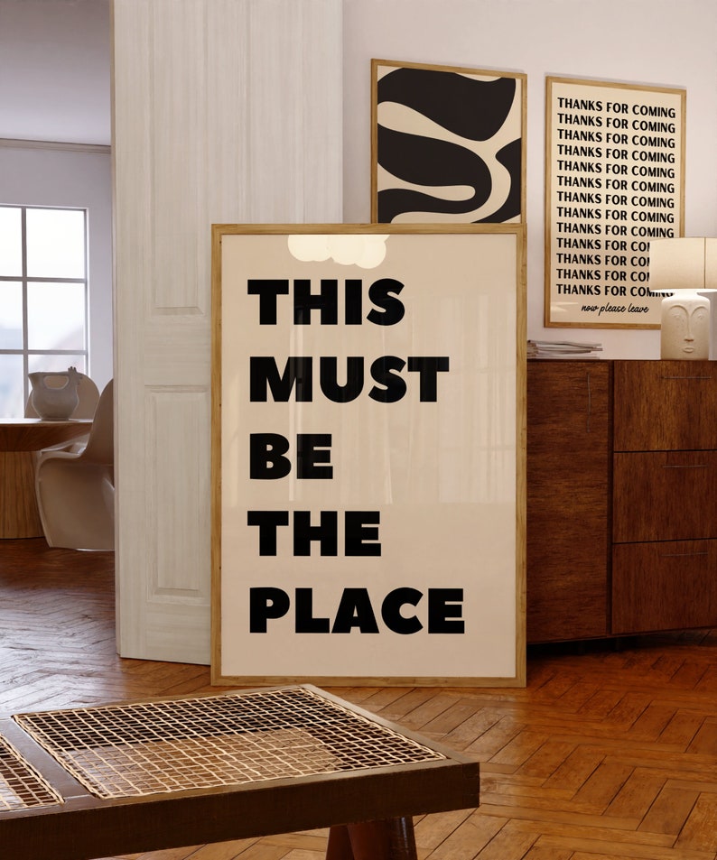 May include: A framed print with the text "THIS MUST BE THE PLACE" in black bold letters on a beige background. The print is hanging on a wall in a room with wooden floors and a wooden cabinet.
