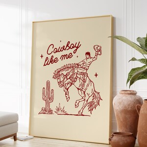 Może przedstawiać: Oprawiony wydruk z czerwoną ilustracją kowboja na koniu, z napisem "Cowboy like me". Przedstawiono r&oacute;wnież kaktus. Grafika ma kremowe tło i złotą ramę.