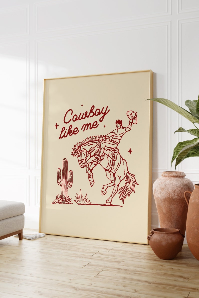 Może przedstawiać: Oprawiony wydruk z czerwoną ilustracją kowboja na koniu, z napisem "Cowboy like me". Przedstawiono r&oacute;wnież kaktus. Grafika ma kremowe tło i złotą ramę.