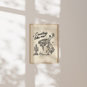 May include: A framed print featuring a black and white illustration of a cowboy riding a bucking bronco. The artwork includes a cactus and the text "Cowboy like me". The print has a beige background and a gold frame.