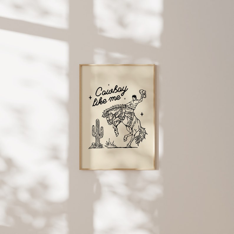 May include: A framed print featuring a black and white illustration of a cowboy riding a bucking bronco. The artwork includes a cactus and the text "Cowboy like me". The print has a beige background and a gold frame.