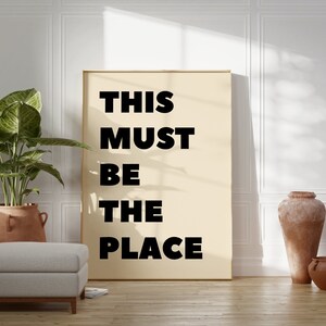 May include: A beige poster with black text that reads "THIS MUST BE THE PLACE" hanging on a white wall in a room with a potted plant, a white ottoman, and two terracotta pots.