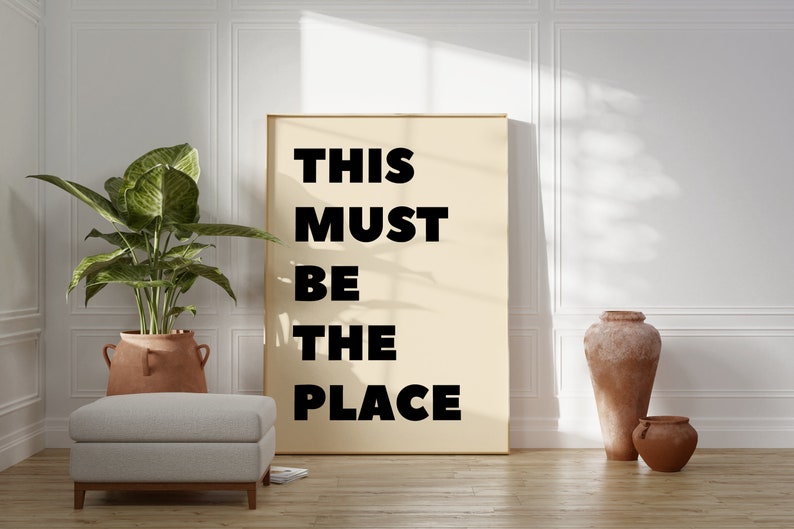 May include: A beige poster with black text that reads "THIS MUST BE THE PLACE" hanging on a white wall in a room with a potted plant, a white ottoman, and two terracotta pots.