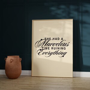 Op de afbeelding: Een ingelijst afdruk met een cr&egrave;mekleurige achtergrond en zwarte tekst die "She had a marvelous time ruining everything" luidt. De afdruk hangt aan een donkerblauwe muur.