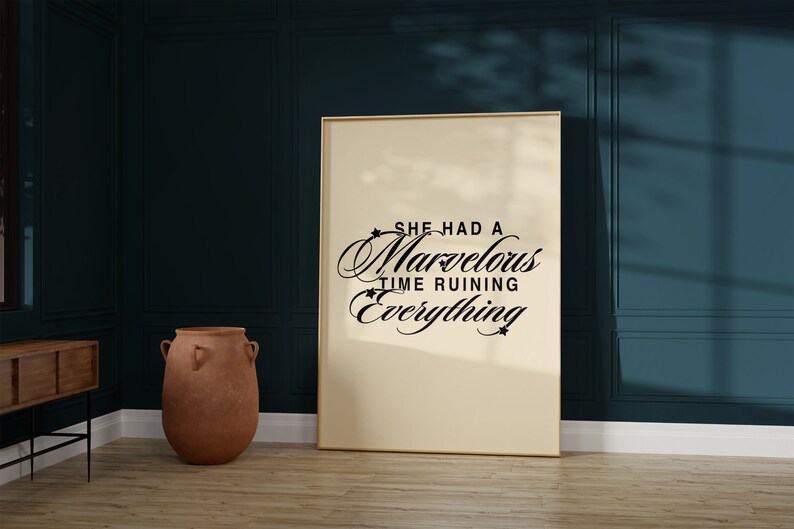 Op de afbeelding: Een ingelijst afdruk met een cr&egrave;mekleurige achtergrond en zwarte tekst die "She had a marvelous time ruining everything" luidt. De afdruk hangt aan een donkerblauwe muur.