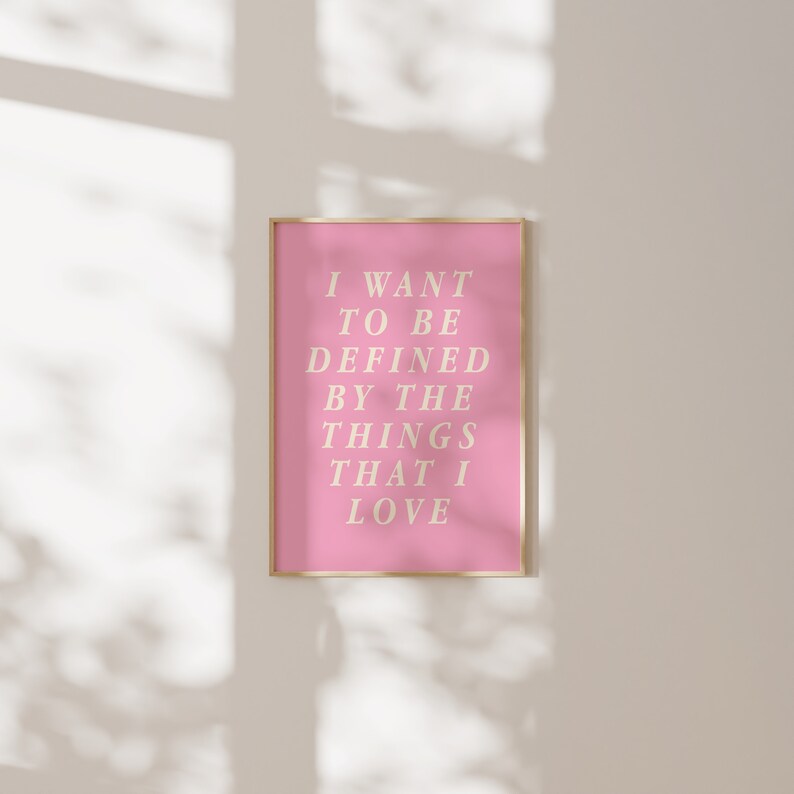 May include: A framed pink art print with the text "I WANT TO BE DEFINED BY THE THINGS THAT I LOVE" in a light beige font. The print has a gold frame and is displayed against a neutral wall.