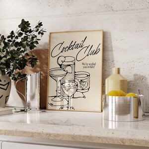 May include: A framed print with the words "Cocktail Club" and the phrase "We're so glad you're here!" in black script. The print features line drawings of cocktail glasses. The print is displayed on a white countertop.