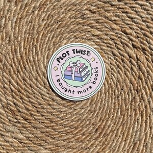Op de afbeelding: Een ronde sticker met de tekst "PLOT TWIST: I bought more books" rond een stapel boeken met een strik. De sticker heeft een lichtroze en groene rand met steraccenten, op een getextureerde, bruine achtergrond.