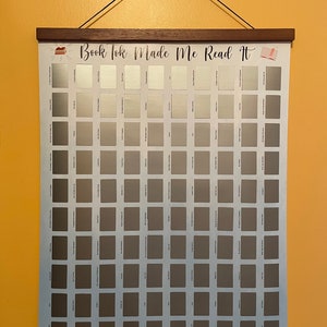 May include: A white poster with a blue background and a grid of 100 grey squares. The poster has the text "Book Tok Made Me Read It" at the top. The poster is hanging on a wall with a wooden frame.