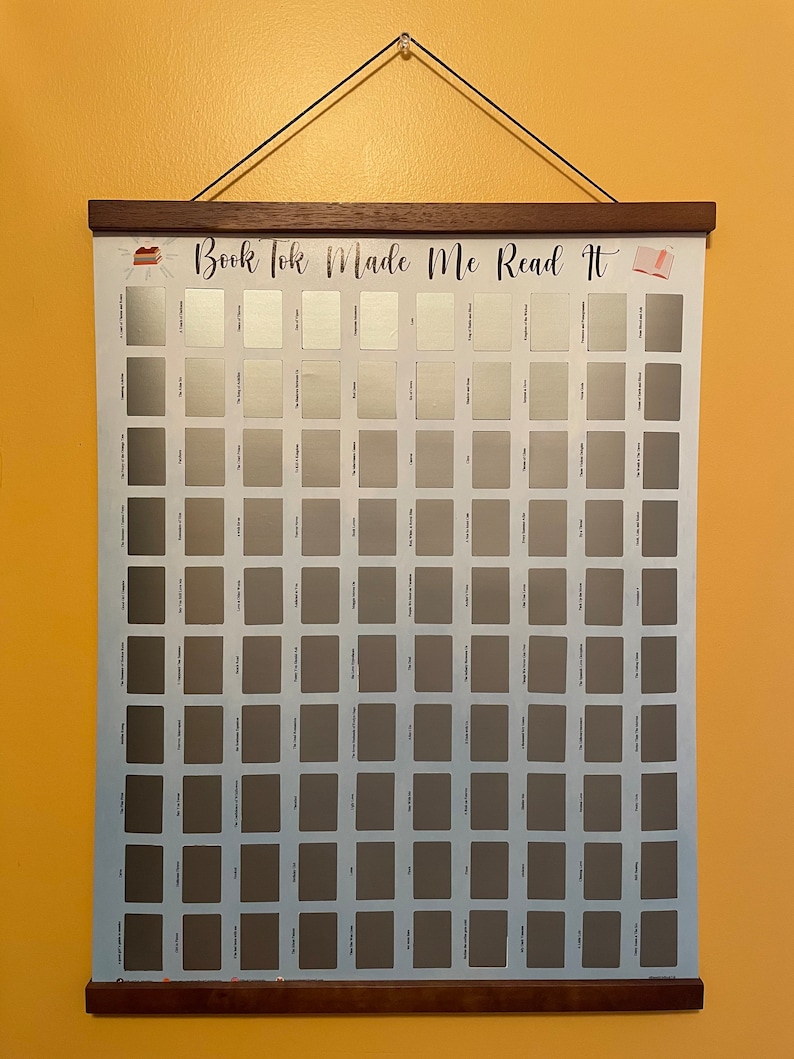 May include: A white poster with a blue background and a grid of 100 grey squares. The poster has the text "Book Tok Made Me Read It" at the top. The poster is hanging on a wall with a wooden frame.