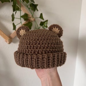 May include: Brown crocheted beanie with brown bear ears. The beanie is made with a soft, textured yarn.