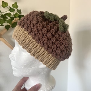 May include: A handmade crochet acorn hat in brown, tan, and green. The hat features a textured, bobble stitch design for the acorn cap and a ribbed brim. The hat is designed to look like an acorn.