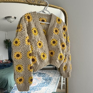 Op de afbeelding: Een gehaakt vest met een zonnebloemmotief. Het vest is beige met gele en bruine accenten. Het heeft een V-hals en lange mouwen.