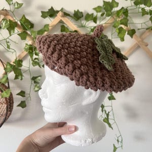 Peut inclure: Béret marron en crochet en forme de gland, avec une tige et deux feuilles vertes. Le chapeau est présenté sur une tête de mannequin blanche. Le béret a une surface texturée et bosselée.