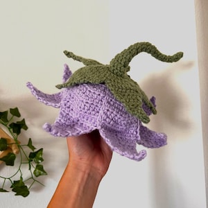 Puede incluir: Una flor de crochet con una campana morada y un tallo verde. La flor se sostiene en una mano.