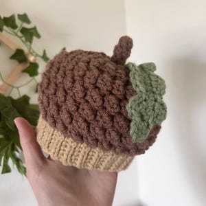 Può includere: Un cappello a forma di ghianda fatto a mano all'uncinetto, marrone e beige, con un dettaglio a forma di foglia verde. Il cappello presenta un punto strutturato, creando un effetto 3D. Il cappuccio della ghianda è marrone e la base è beige.
