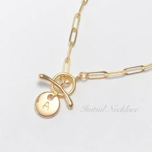 May include: A gold chain necklace with a toggle clasp and a round pendant with the letter "A" engraved on it. The necklace is on a white background. "Initial Necklace" is written below the necklace.