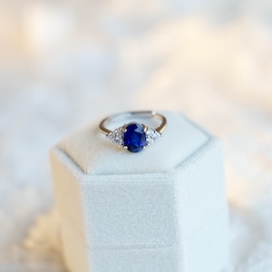 May include: A silver ring with a large oval blue sapphire gemstone and smaller clear gemstones on the band.