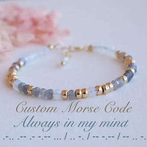 May include: A delicate bracelet made with grey and white beads and gold accents. The bracelet is a custom Morse code design that spells out "Always in my mind".